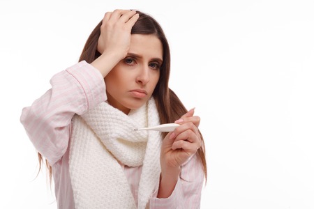 Young sick woman in scarf and pajamas looking at thermometer. Sick girl with a thermometer on a white background, isolate, flu, coldsの写真素材