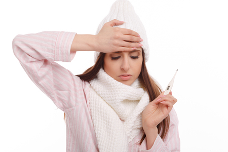 Young sick woman in scarf and pajamas looking at thermometer. Sick girl with a thermometer on a white background, isolate, flu, coldsの写真素材