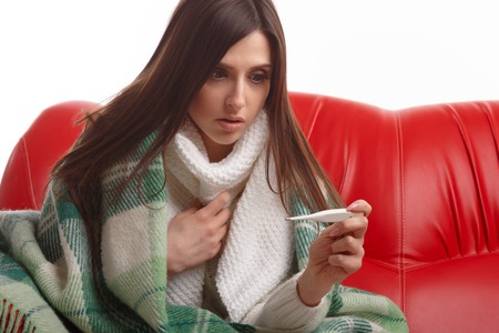 Young sick woman in scarf and pajamas looking at thermometer. Sick girl with a thermometer on a white background, isolate, flu, coldsの写真素材