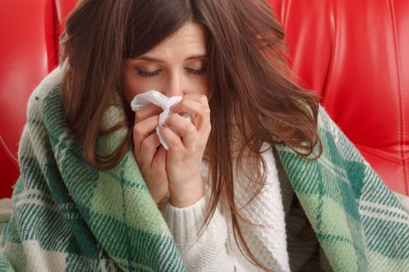 Sick young woman sitting on a red couch, and blowing his nose into a napkin. Woman sick blowing nose isolated. Sick Woman. Flu. Woman Caught Coldの写真素材