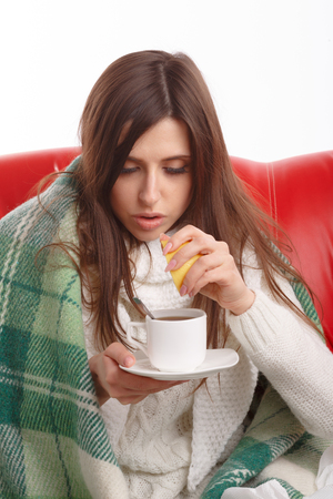 Sick young woman sitting on a red sofa and drinking tea with lemon. Young female caught cold, feeling bad, wrapped up in blanket, squeezing lemon to her tea.の写真素材