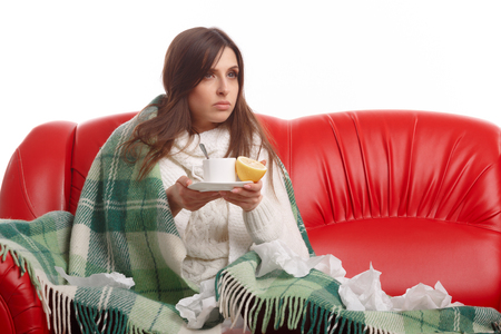 Sick young woman sitting on a red sofa and drinking tea with lemon. Young female caught cold, feeling bad, wrapped up in blanket, squeezing lemon to her tea.の写真素材