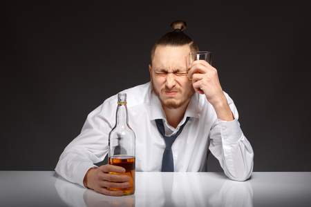 A young man in a white shirt sits at a table and suffers from a hangover Addicted to alcohol, alcoholism concept, social problemの写真素材