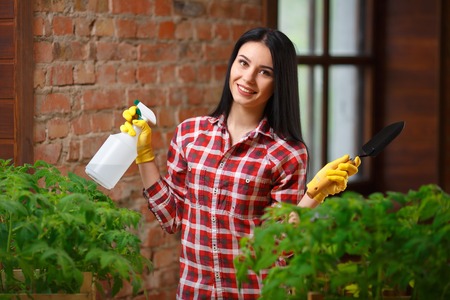 Gardening is her passion. Charming young gardener posing indoors holding shovel and water spray looking to the camera smiling happily.の写真素材