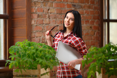 Looking closer. Portrait of a beautiful young woman posing indoors holding a laptop looking at her tomato seedlings on the table.の写真素材