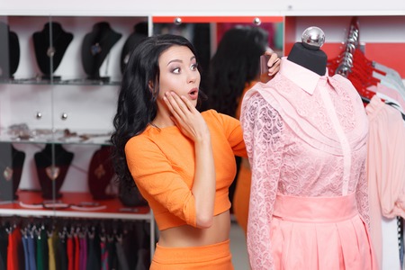 Young woman shopper astonished price on the dress. Young woman shopping in a clothing store. Caucasian shopper girl choosing pink dress in shop during sale. Woman shopping for dress. Fashion shoppingの写真素材