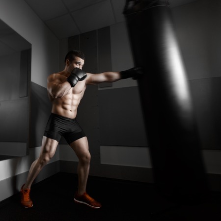 Male boxer training with punching bag in dark sports hall. Young boxer training on punching bag. Male boxer as exercise for the big fight. Boxer hits punching bag. Young masculine male athlete.の写真素材
