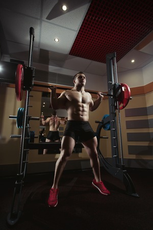 Bodybuilder squatting with a barbell. Bodybuilder working with barbell. Fitness man doing squats with the barbell at the gym. Smith Machine in the gym. Fitness man in the gym. Perfect healthy figureの写真素材