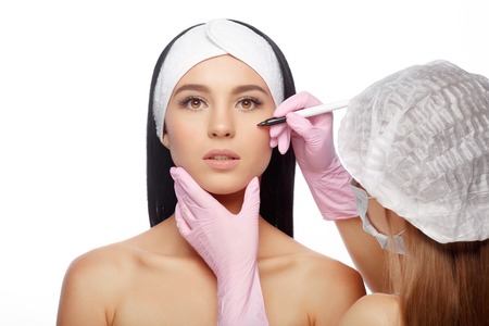 Young Woman before Plastic Surgery Operation Cosmetology. Doctor does line on a female face before plastic surgery. Beautician touching woman face. Beauty Face. Isolate on white backgroundの写真素材