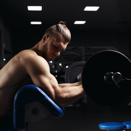 Bodybuilder in the gym. Muscular man working out doing exercises with barbell at biceps, strong male naked torso in the gym. Handsome bodybuilder with big muscles, posing at the camera in the gymの写真素材