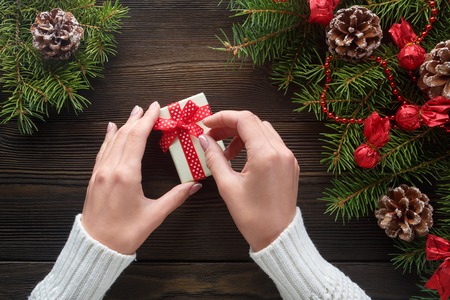 Beautiful female hands holding a Christmas present in box with red bow. Christmas gift on a background of wooden table and Christmas tree.の写真素材