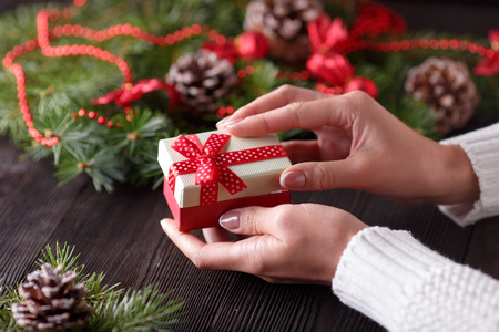 Beautiful female hands holding a Christmas present in box with red bow. Christmas gift on a background of wooden table and Christmas tree.の写真素材
