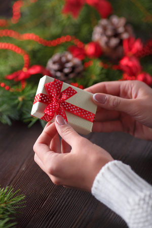 Beautiful female hands holding a Christmas present in box with red bow. Christmas gift on a background of wooden table and Christmas tree.の写真素材