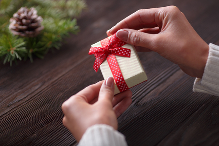 Beautiful female hands holding a Christmas present in box with red bow. Christmas gift on a background of wooden table and Christmas tree.の写真素材