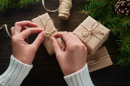 Beautiful female hands are packed Christmas gift in brown  paper, rope. Gift box in brown  paper tied with string. Christmas gift in  paper with decoration on rustic wooden background.の写真素材
