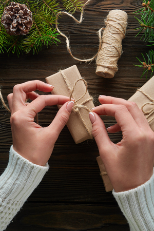 Beautiful female hands are packed Christmas gift in brown  paper, rope. Gift box in brown   paper tied with string. Christmas gift in  paper with decoration on rustic wooden background.の写真素材