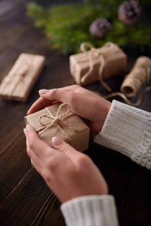 Beautiful female hands are packed Christmas gift in brown  paper, rope. Gift box in brown  paper tied with string. Christmas gift in  aft paper with decoration on rustic wooden background.の写真素材