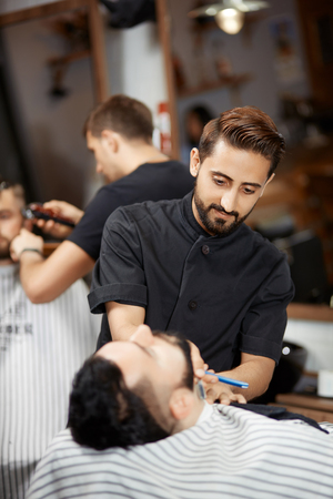 Handsome hairstylist in barber shop cutting bread with razor for brunet man.の写真素材