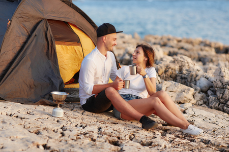 Happy smiling couple sitting face to face at rocky beach on near tent.の写真素材