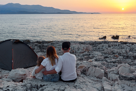 Back view of family, sitting on rock beach, admiring setting sun over sea.の写真素材