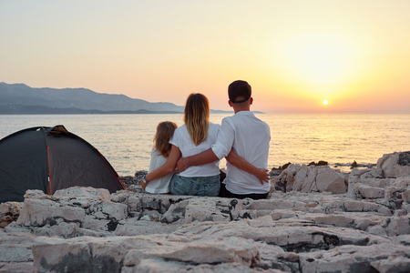 Back view of cute young family, sitting on rock beach near tent and admiring setting sun over sea. Mom, dad and little daughter embracing and having great time together. Concept of friendly family.の写真素材