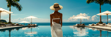 A woman dressed in a white dress and straw hat stands confidently in front of a pristine swimming pool at a resort setting.の素材