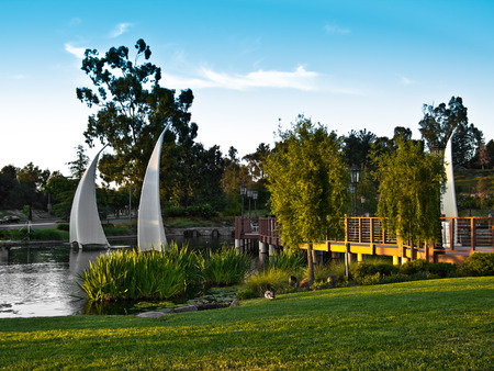 Late afternoon sun throws long shadows across the lawn and a footbridge at a park pond の写真素材