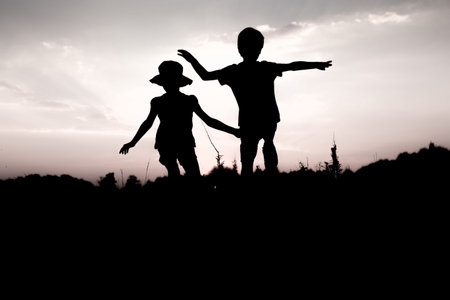 Silhouettes of kids jumping off a cliff at sunset. Little boy and girl jump raising hands up high. Brother and sister having fun in summer. Friendship, freedom concept. Fraternal twins on vacation in mountains.の写真素材