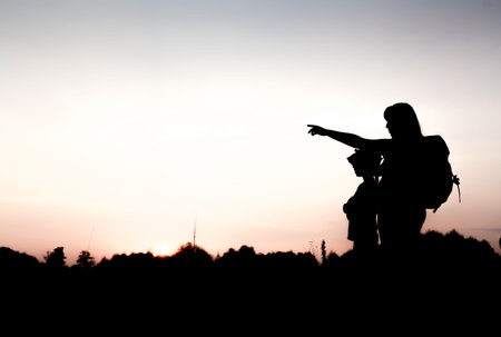 Silhouettes of mother and child hiking at sunset. Mom with backpack pointing at something distant....