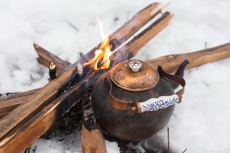 Copper kettle over an open fire in winter. Boiling kettle on firewood. Open fire cooking. Snow around. Lifestyle, camping. Blurred background.の写真素材
