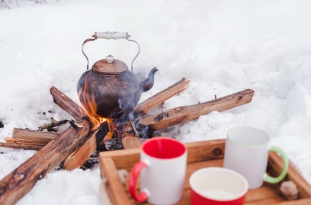 Romantic winter picnic. Two cups and a bowl on a wooden tray in snow. Copper kettle over an open fire on background, blurred. Boiling kettle on firewood. Lifestyle, camping.の写真素材