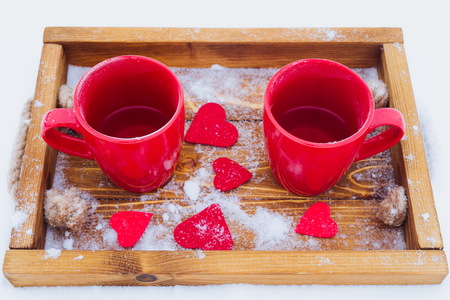 Two red cups with tea on a wooden tray in snow. Romantic Valentine's day winter picnic, hot tea, couple. Lifestyle, time together outdoors, Valentine's day. Snowflakes, hoarfrost and heart shape on a trayの写真素材
