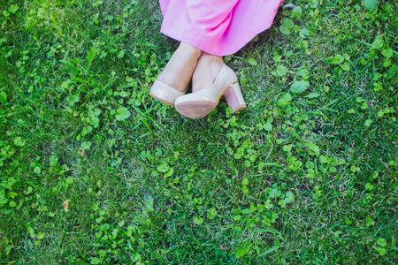 Feet of a bride on a green grass background. Happy bride in simple pink dress and stylish beige shoes sitting on a grass on wedding day. Summer wedding outdoors, happy people, copy space.の写真素材
