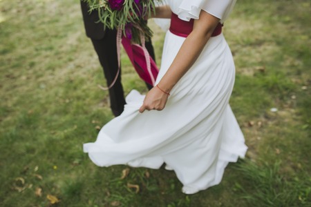 Happy couple on a walk on their wedding day. Bride in a stylish white dress dancing for a groom outside. People in love. Dress flying up in wind. Beautiful wedding bouquet, nice flowers. Blurred.の写真素材