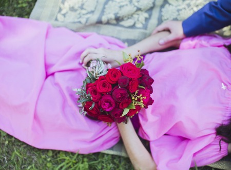 Happy couple in love lying in green grass on their wedding day. Bride in simple stylish pink dress holds beautiful wedding bouquet, nice flowers. Happy people on summer day outdoors.の写真素材