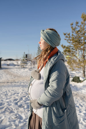 A happy pregnant young woman with her eyes closed, hugging her stomach in the park in winter, in a warm down jacket.の写真素材