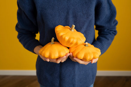 Hands of a young caucasian woman holding pumpkins against a yellow wall