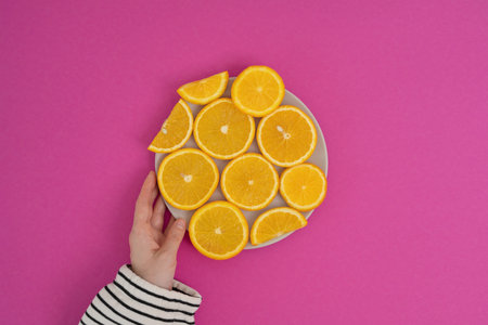 Female hand holding a plate with slices of orange on a pink backgroundの写真素材