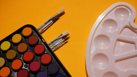 Palette of watercolors and brushes on a yellow background.