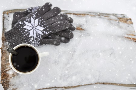 A white Cup of coffee and knitted gray gloves with a pattern on a wooden bench in the snow during a snowfall. Cozy winter card.の写真素材