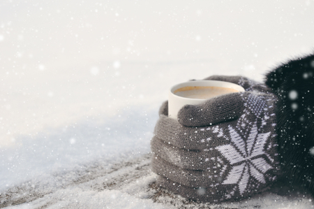 Winter picture: hands in knitted gray gloves holding a Cup of hot coffee on a snowy day on a wooden rustic background in the village. copyspace.の写真素材