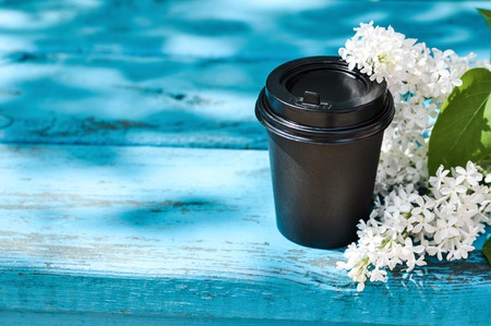 Black cardboard Cup of coffee and a branch of lilac on a blue wooden bench against green grass and sunlight. Mockup. Copy spaceの写真素材