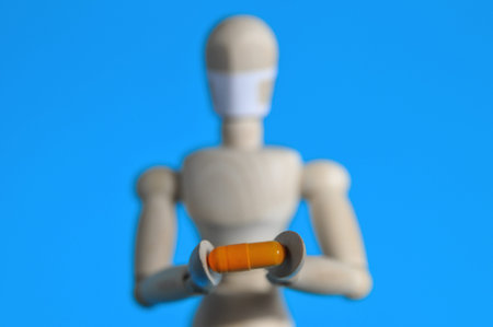 A wooden figure in a medical mask holds an orange pill on a blue background. Coronavirus, pandemic and epidemic conceptの写真素材