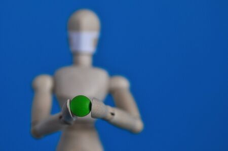 Wooden figure of a mannequin in a medical mask holding a green pill on a blue background. Coronavirus, pandemic and epidemic conceptの写真素材