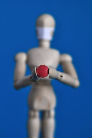 Wooden figure of a mannequin in a medical mask holding a red pill on a blue background. Coronavirus, pandemic and epidemic conceptの写真素材