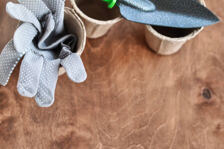 Grey reusable gardening gloves in a peat eco pot for seedlings and a metal shovel next to it on a brown wooden background. copy spaceの写真素材