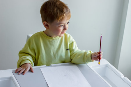 Cheerful little boy in a yellow sweater draws with colored pencils on paper at a tableの写真素材