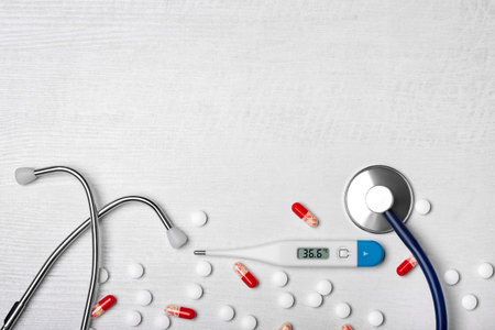 Stethoscope, electronic body thermometer and scattered pills on a light wooden background. Top view....
