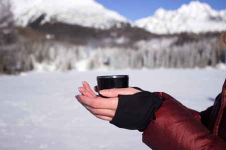 A young woman holds a cup of tea against the background of mountains in winter. Strbske Pleso Lake, Slovakiaの写真素材
