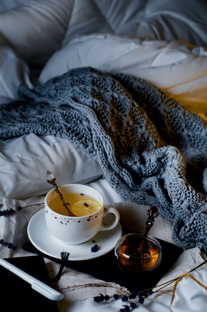 Evening tea in bed with gentle lighting. Tea with lemon and honey on a tray and knit plaid on a white bedの写真素材